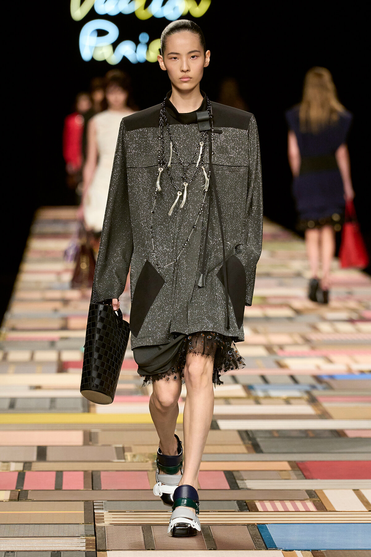 Model walks a colorful tiled runway in a glittery gray oversized coat with black patches, layered necklaces, and a black textured bag.
