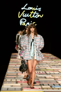 Model walks the Louis Vuitton runway wearing a oversized striped gray jacket with gathered shoulders, mint scarf, and brown sandals, carrying a black handbag against a neon Louis Vuitton Paris backdrop.