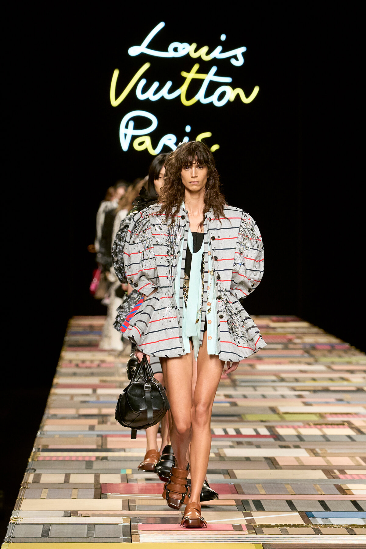 Model walks the Louis Vuitton runway wearing a oversized striped gray jacket with gathered shoulders, mint scarf, and brown sandals, carrying a black handbag against a neon Louis Vuitton Paris backdrop.