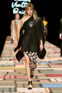 Model walks the runway in a black pleated, one-shoulder top over a striped wrap skirt, paired with chunky sandals, on a multicolor tiled catwalk with blurred audience in the background.