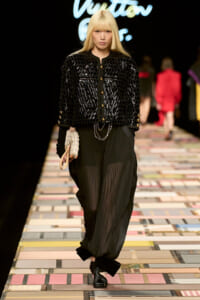 Runway model wearing a black textured quilted jacket with gold buttons, sheer black pants, and a small beige clutch.