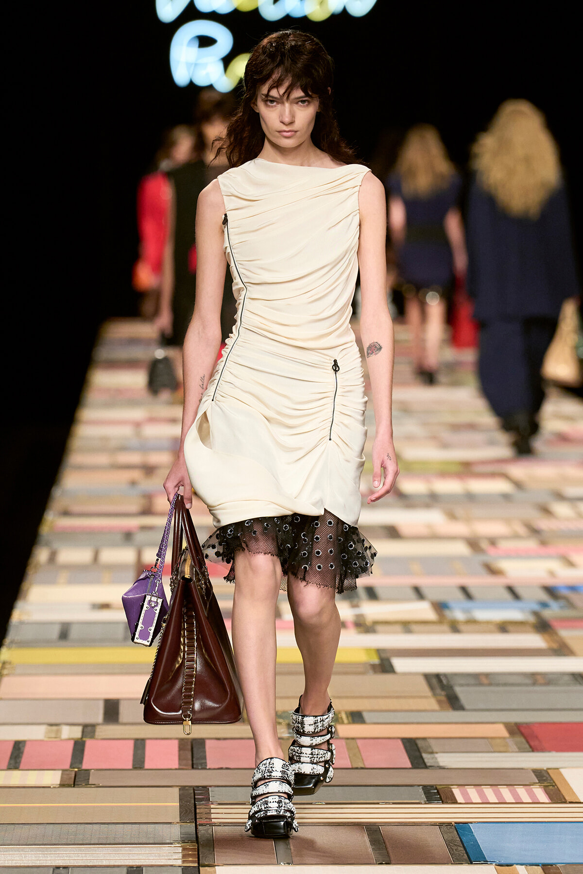 Model walking a fashion show runway in a cream draped dress with a black lace hem, carrying a large brown handbag and a smaller purple bag nearby.