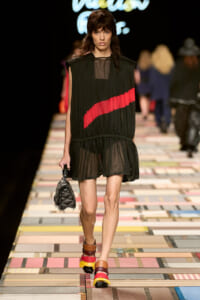 Model walks the runway in a sheer black sleeveless dress with a red diagonal stripe, carrying a small fringed handbag and wearing multicolored platform sandals on a tiled runway