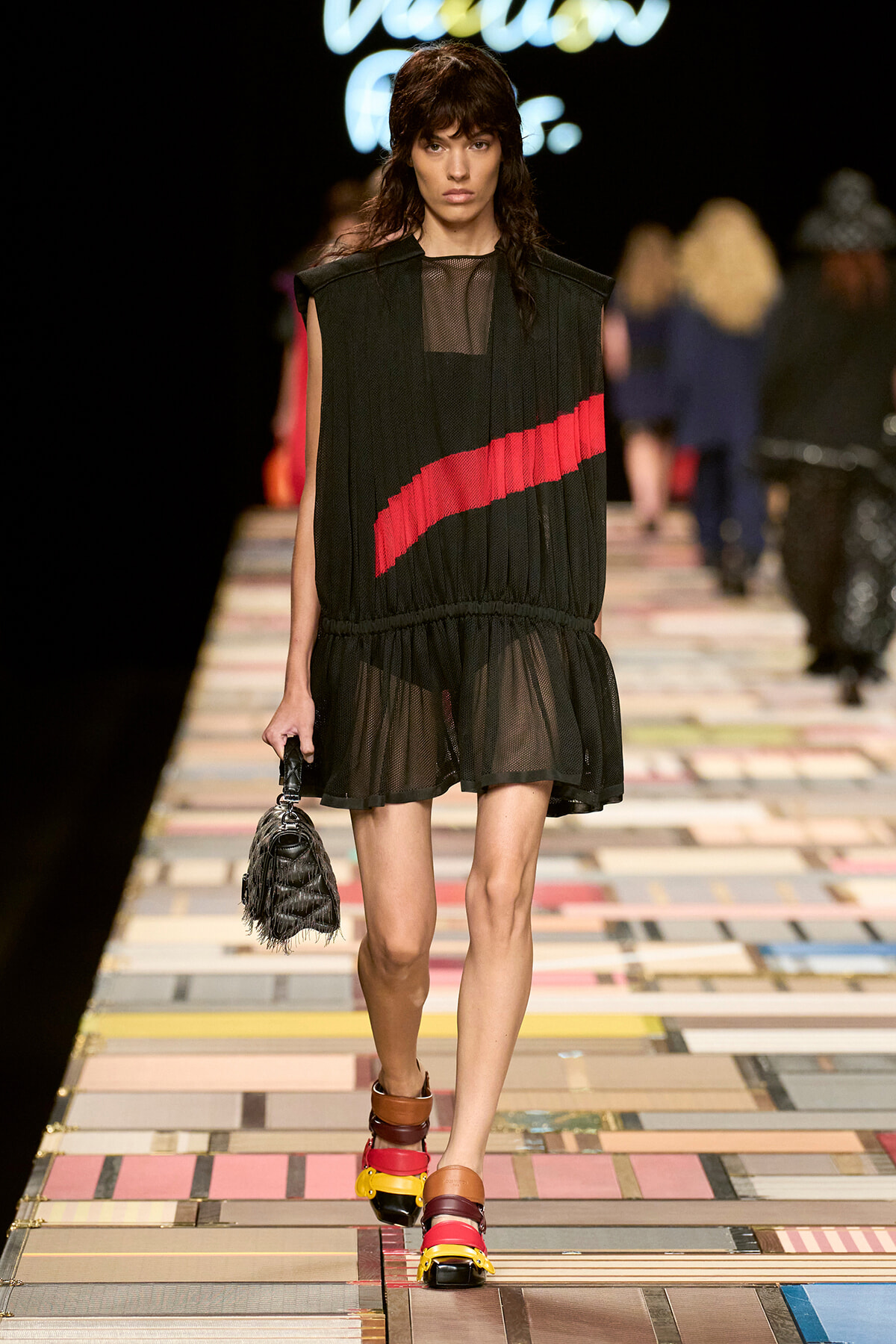Model walks the runway in a sheer black sleeveless dress with a red diagonal stripe, carrying a small fringed handbag and wearing multicolored platform sandals on a tiled runway