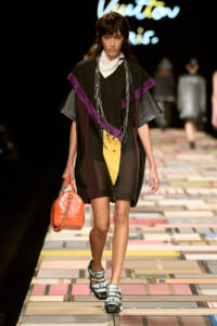 Model walking a fashion runway in a loose black outfit with purple accents, yellow panel, chunky sandals, and an orange crocodile-embossed bag.