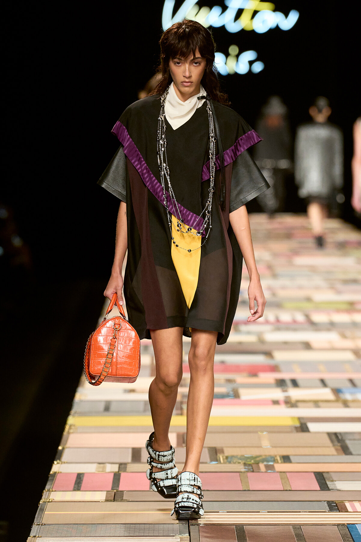 Model walking a fashion runway in a loose black outfit with purple accents, yellow panel, chunky sandals, and an orange crocodile-embossed bag.