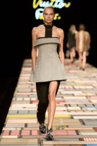 Runway model walking the catwalk in a gray off‑the‑shoulder dress layered over black pants, with a black neck strap and patterned black‑and‑silver sandals.