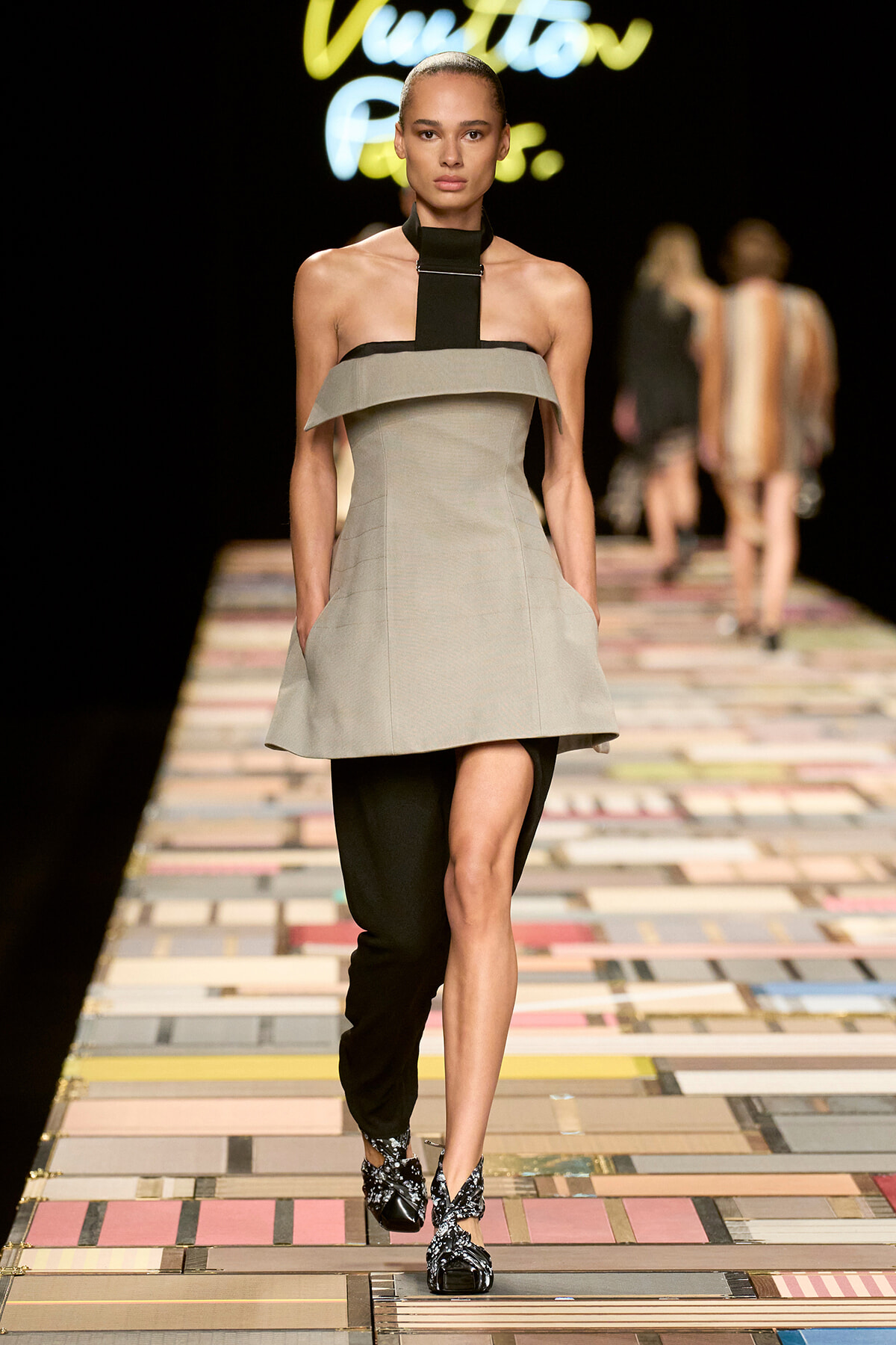 Runway model walking the catwalk in a gray off‑the‑shoulder dress layered over black pants, with a black neck strap and patterned black‑and‑silver sandals.