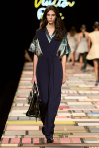 Model walks a dark runway wearing a navy jumpsuit with metallic green sleeves, carrying a black handbag.