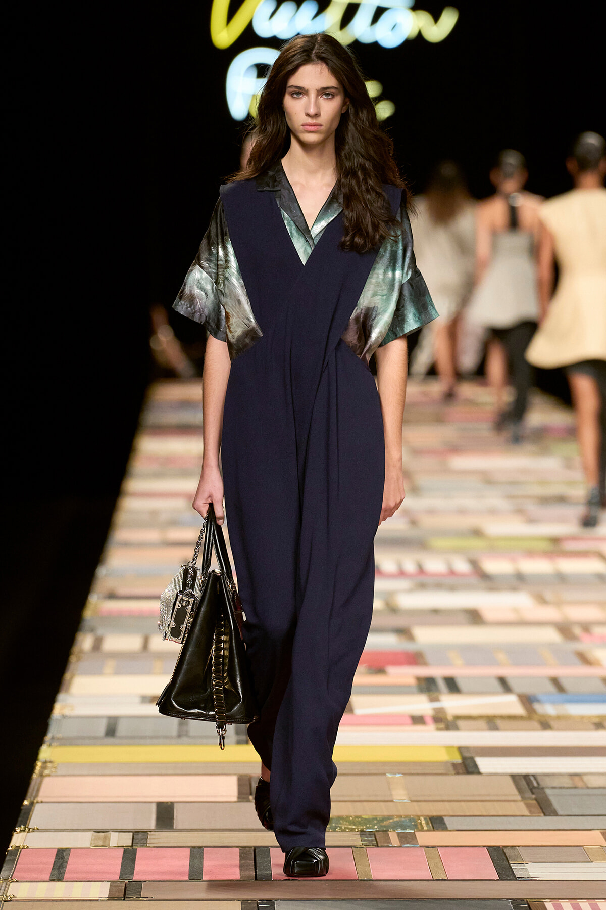 Model walks a dark runway wearing a navy jumpsuit with metallic green sleeves, carrying a black handbag.