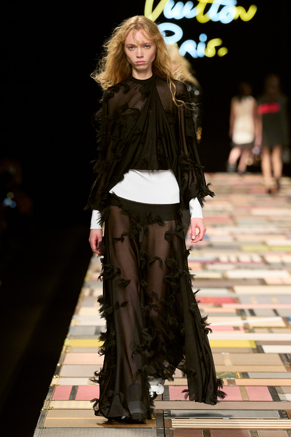 Model walks a fashion runway in a sheer black, ruffle-adorned gown over a white underlay on a colorful patchwork catwalk.