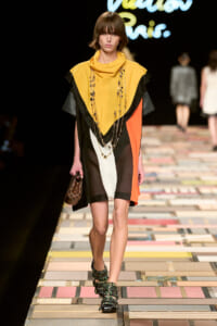 Model walking the runway in a color-block dress: mustard yellow, black, white, and orange panels with a draped neckline and layered necklaces; she carries a small checkered handbag and wears multi-strap, embellished sandals.