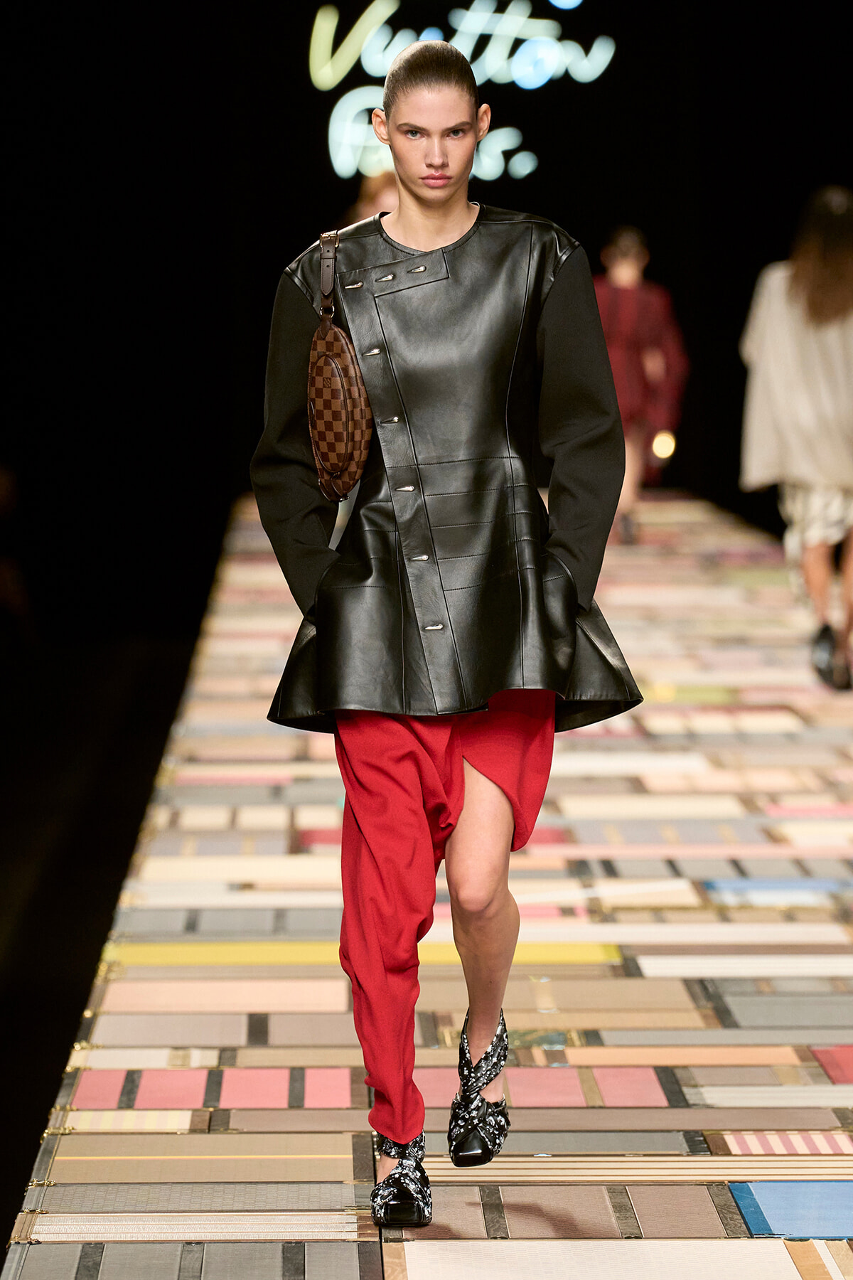 Model walking a runway in a black leather tunic with red asymmetrical pants, a brown checkered bag slung over the shoulder, and embellished sandals.