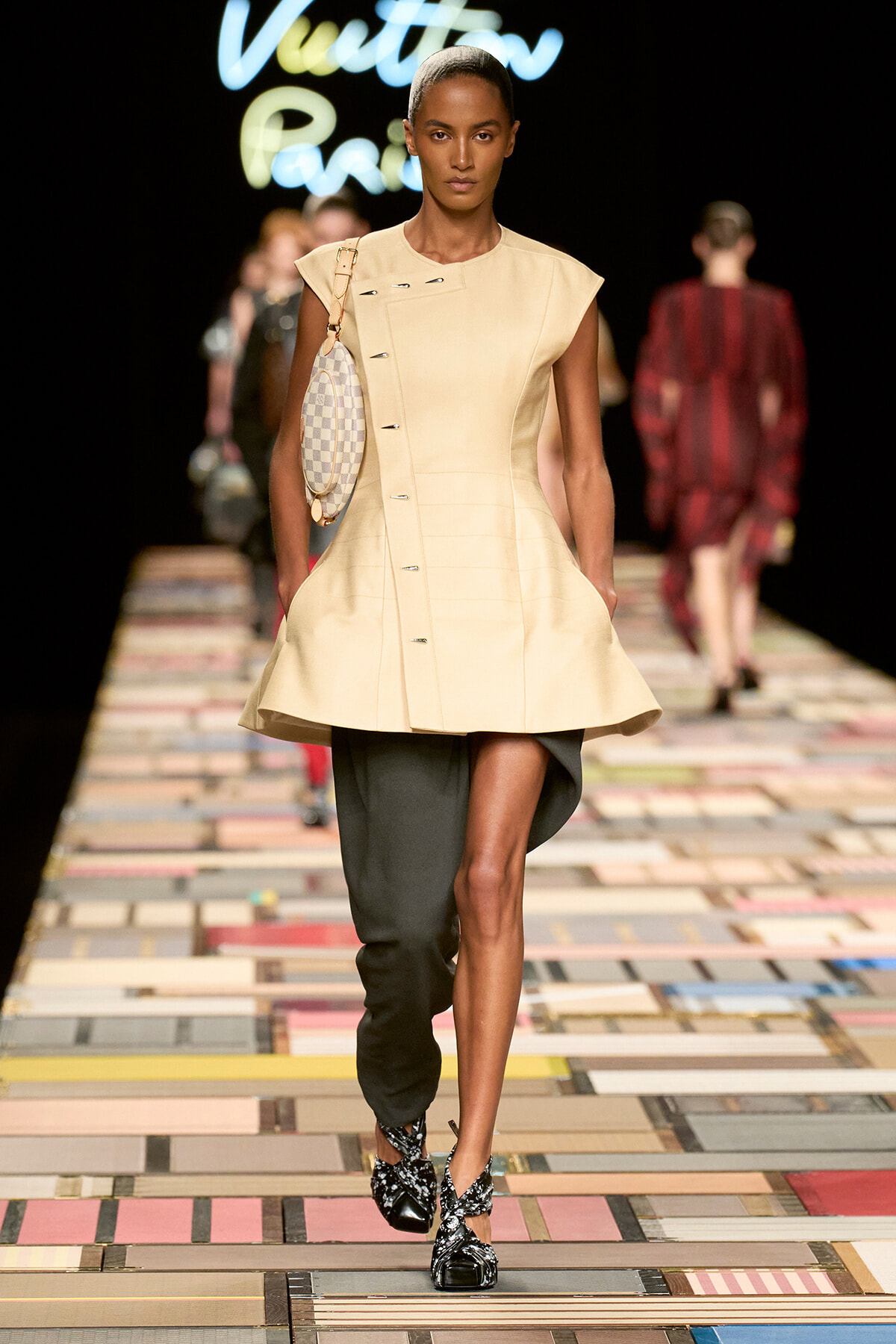Model walks a fashion runway in a cream sleeveless coat with an asymmetric button line, paired with black draped pants and a checkered oval handbag over the shoulder.