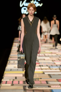 Model walks the runway in a dark gray sleeveless jumpsuit, carrying a vintage metal suitcase.