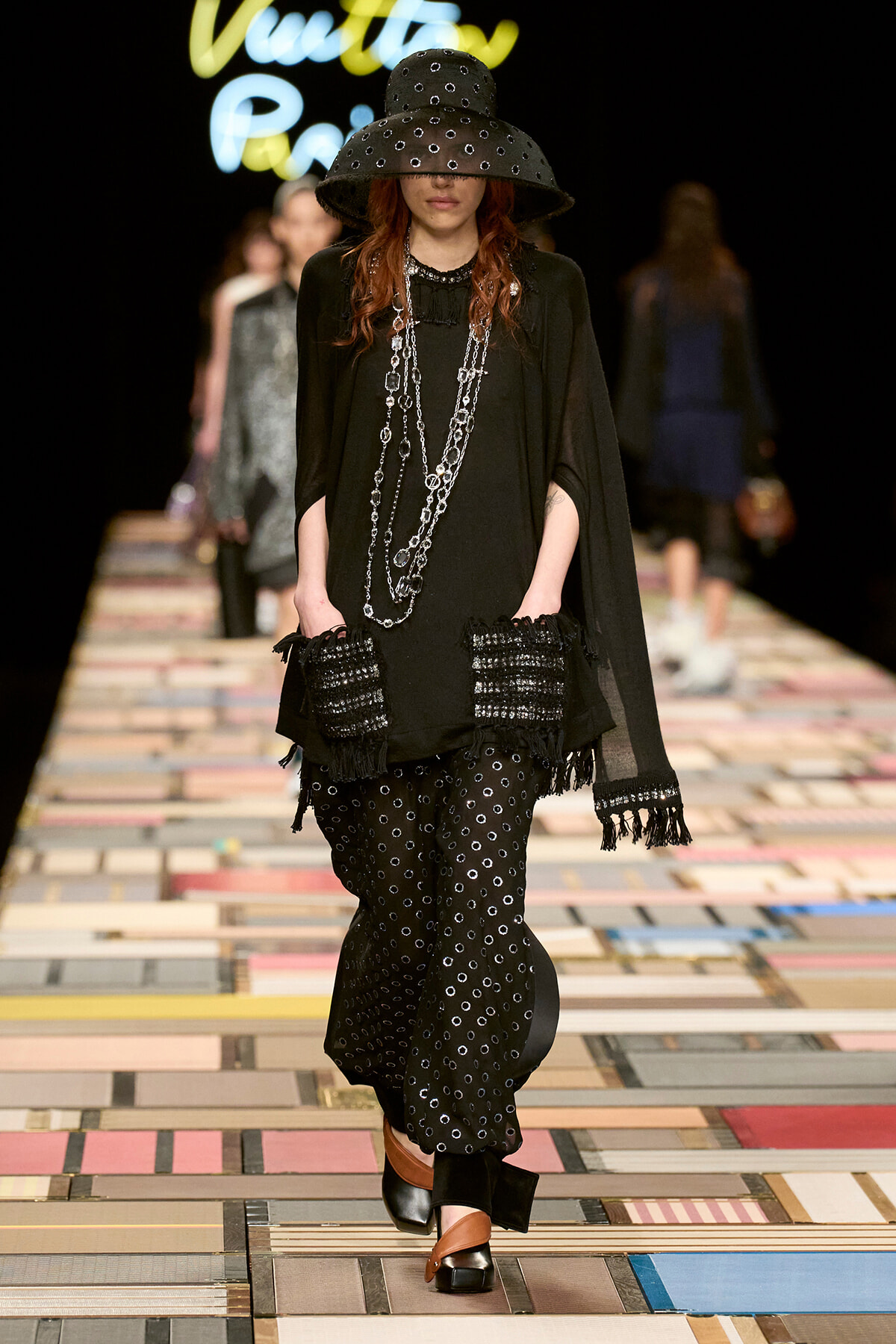 Model walks the runway in a black ensemble with an oversized studded hat, layered silver chains, fringe gloves, and dotted wide-leg pants in a fashion-show look.
