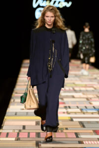Fashion runway model walks the catwalk in a navy cape-style top and matching pants, carrying beige tote and a small green bag on a colorful tiled runway.