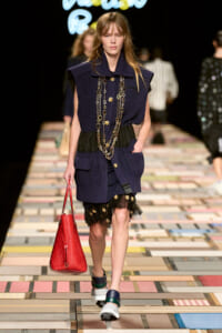 Model on a runway wearing a navy sleeveless coat with gold buttons, layered chains, and a red textured handbag