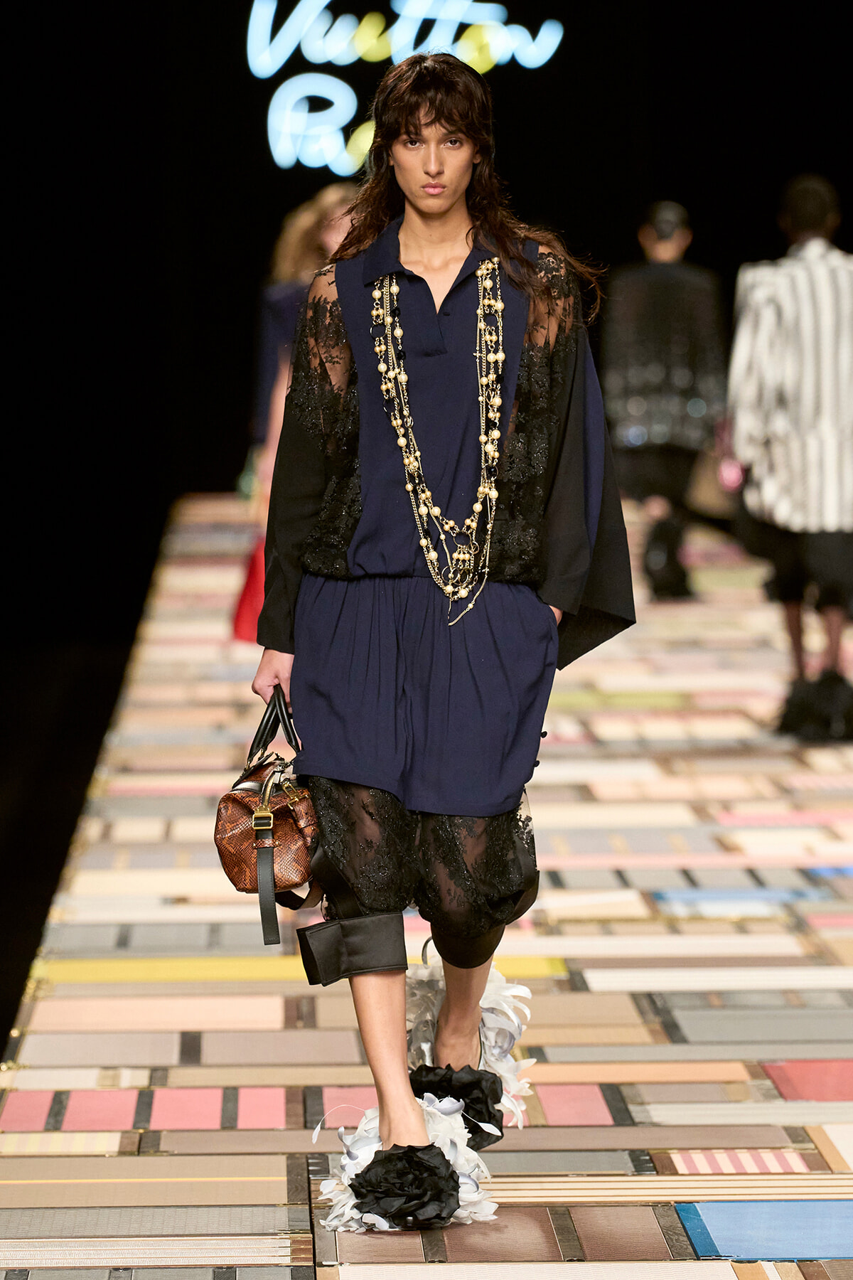 Model walks a fashion show in a navy outfit with lace sleeves, layered jewelry, and a brown handbag; bold floral foot embellishments visible on the runway.