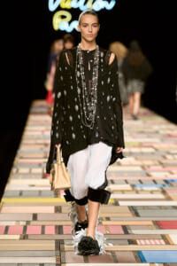 Model walks the runway wearing a black polka-dot sheer top, white draped pants, layered silver necklaces, and a beige handbag.
