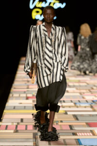 Model walks the runway in a black-and-white striped oversized blazer with puff shoulders, black cropped trousers, and feathered black footwear, carrying a brown clutch.