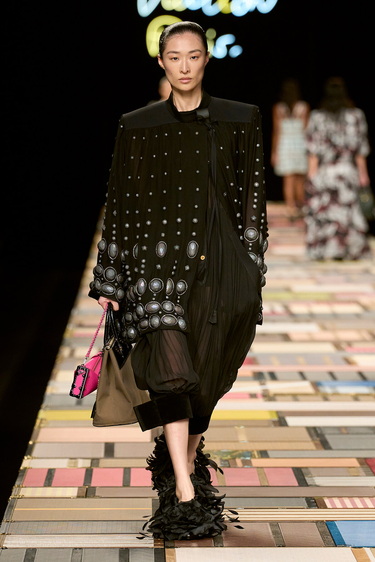 Model walking a runway in a black studded cape with circular metallic embellishments, paired with sheer black pants and feathered shoes, carrying a pink handbag on a multicolored tiled catwalk.
