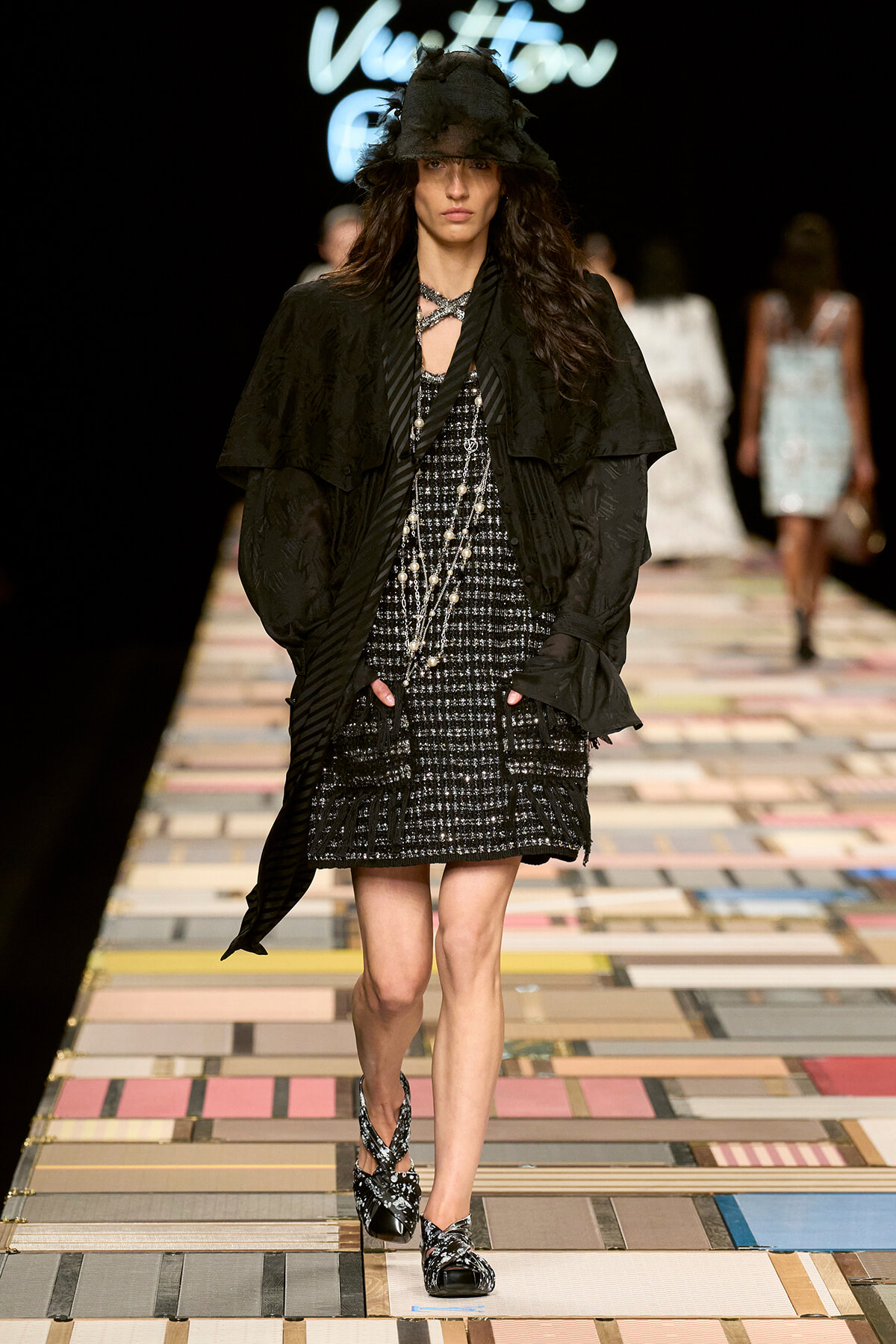 Runway model in black layered coat over a tweed black-and-white dress with pearl necklace, hat, and patterned heels walking a colorful tiled runway