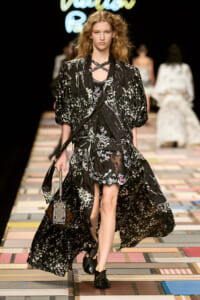 Model walks a runway in a black and white floral-print cape over a sheer lace dress, with a cross-neck necklace and braided black sandals, carrying a small handbag.