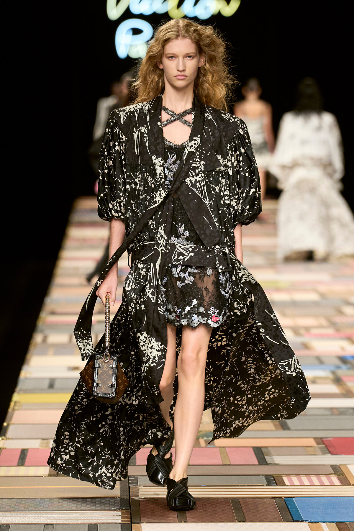 Model walks a runway in a black and white floral-print cape over a sheer lace dress, with a cross-neck necklace and braided black sandals, carrying a small handbag.