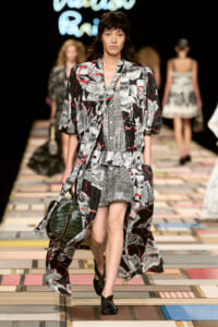 Model walks the runway in a black, white, and red abstract-print coat over a matching patterned dress; carries a dark green crocodile-embossed bag and wears black twisted sandals, with other models in the background.