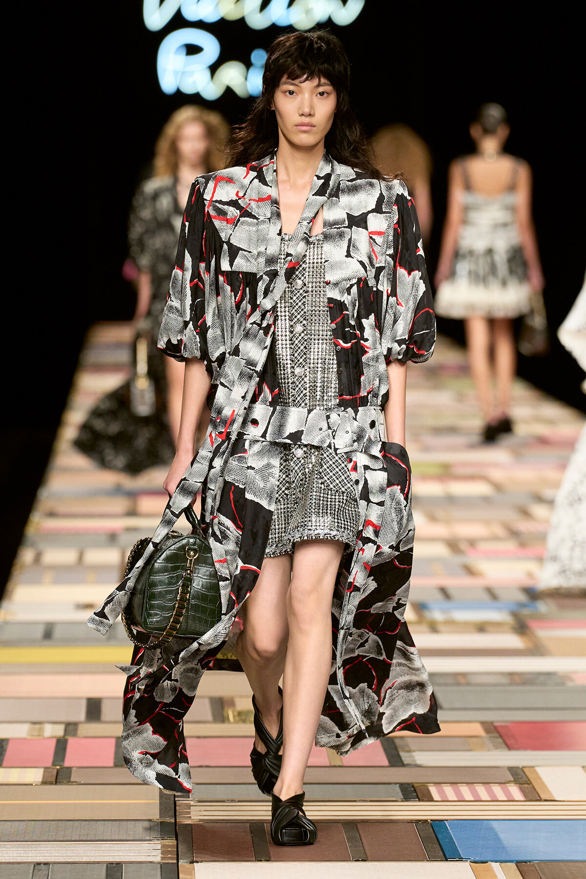 Model walks the runway in a black, white, and red abstract-print coat over a matching patterned dress; carries a dark green crocodile-embossed bag and wears black twisted sandals, with other models in the background.