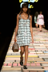 Model walking a fashion runway in a light blue tweed slip dress with floral embellishments, carrying a small brown Louis Vuitton handbag and wearing black twisted flats.