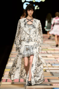 Runway model wearing a cream floral-print trench with a belt over a metallic patterned mini dress, walking the catwalk.
