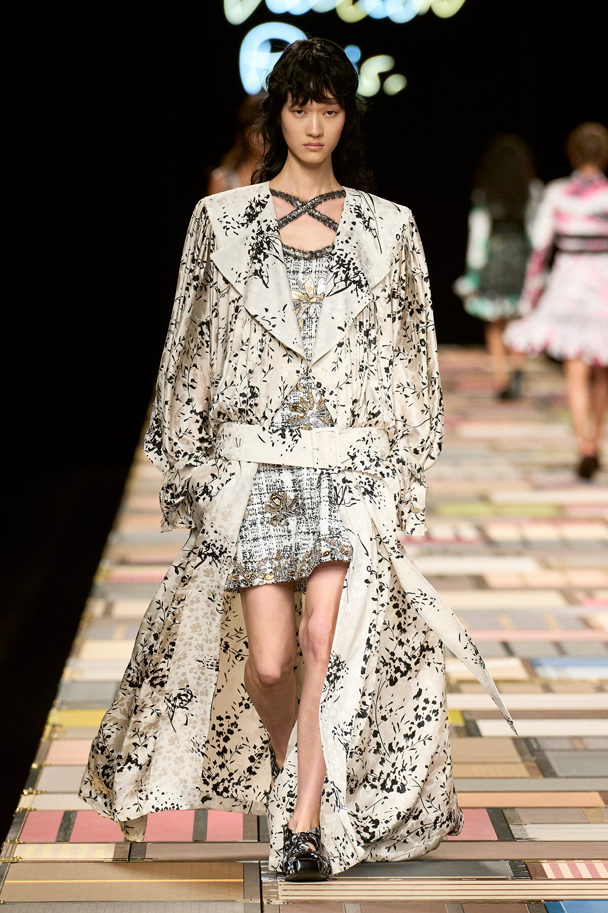 Runway model wearing a cream floral-print trench with a belt over a metallic patterned mini dress, walking the catwalk.