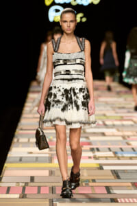 Model walking the runway in a black-and-white tiered dress with metallic straps, carrying a small brown handbag.