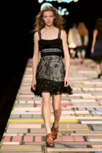 Model walks the runway in a black, tiered mini dress with circular stud embellishments and ruffled hem, paired with brown strappy sandals.