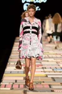 Model walks the runway in a pink, white, and black patterned dress with belt-like bands and a ruffled hem, carrying a small brown purse.
