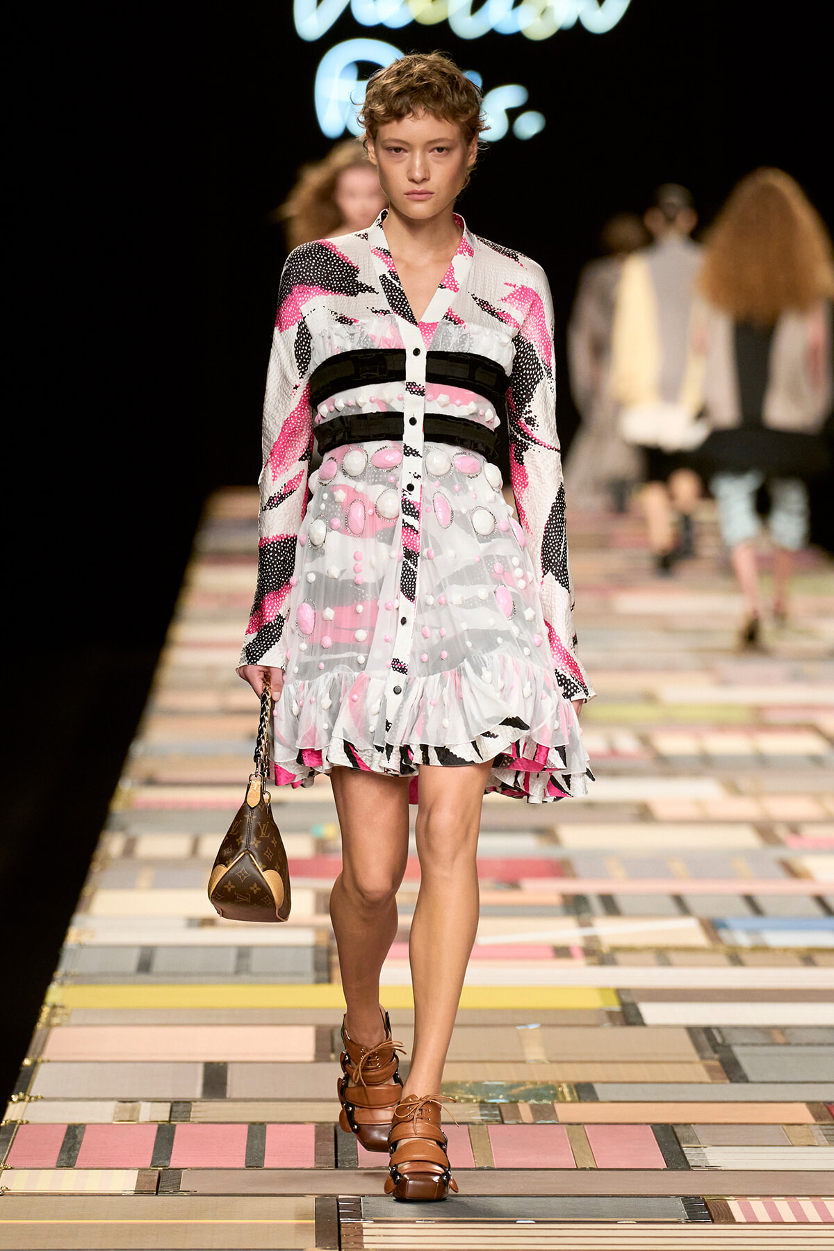 Model walks the runway in a pink, white, and black patterned dress with belt-like bands and a ruffled hem, carrying a small brown purse.