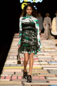 Model walking a fashion show in a green, black, and white patterned dress with ruffles and a wide belt, carrying a small brown handbag.