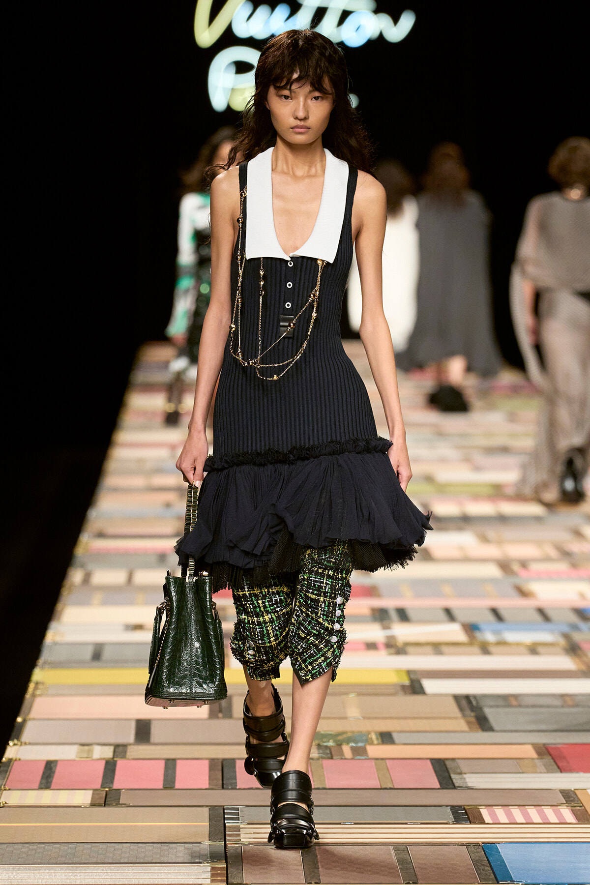 Model walks a fashion show wearing a navy sleeveless knit dress with a white collar, layered gold chains, and a frilled hem over plaid green pants; carries a dark green handbag and wears black sandals.