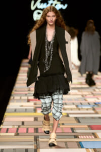 Model walks a fashion runway wearing a black layered top with a beige cape-style jacket, long silver chains, a ruffled black skirt, plaid cropped pants, and beige strappy sandals.