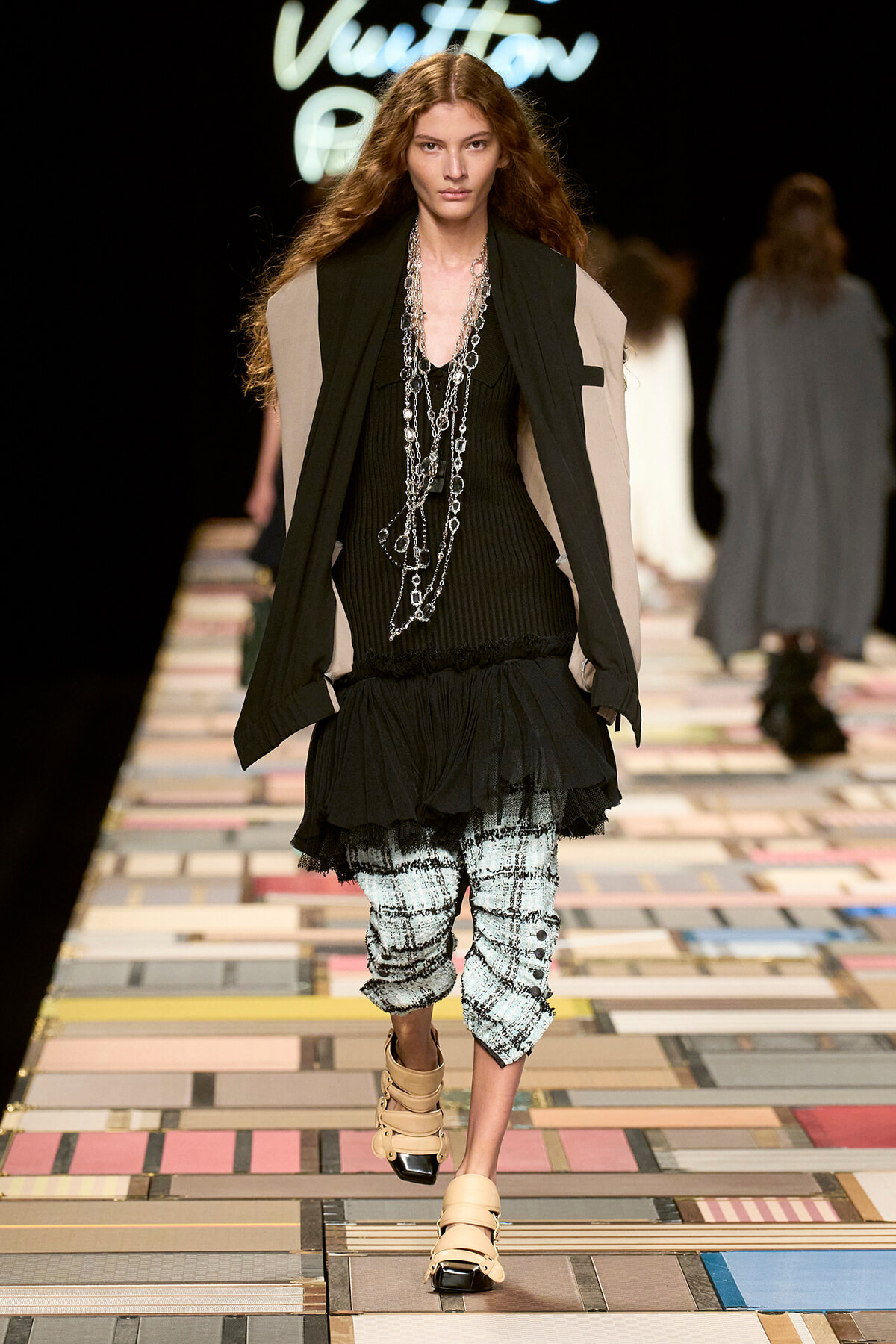Model walks a fashion runway wearing a black layered top with a beige cape-style jacket, long silver chains, a ruffled black skirt, plaid cropped pants, and beige strappy sandals.
