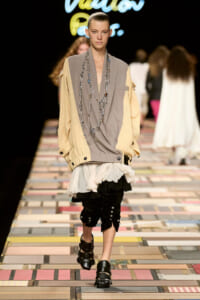 Model on a runway wearing a beige-gray draped top, layered ruffled skirt over black cropped pants with metal studs, and chunky black sandals; long silver necklaces cascading down the front.