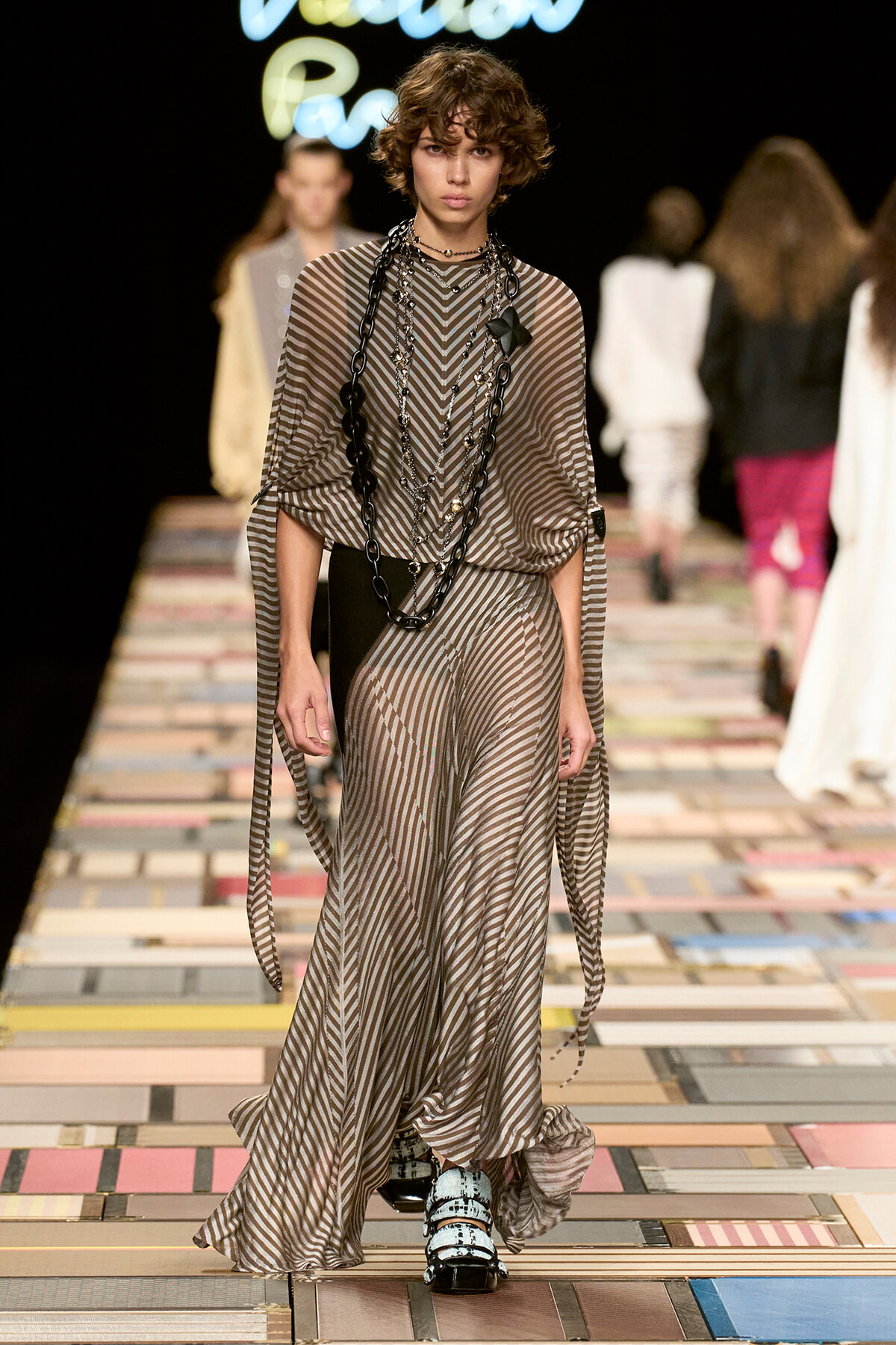 Model walks the runway in a brown and beige chevron-striped gown with long draped sleeves and layered chains as accessories.