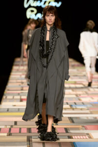 Model walks the runway in a voluminous gray draped coat with long black chain necklaces and black feathered shoes on a patterned catwalk surface.