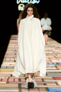Model on a fashion runway in an off-white, cape-style coat with a high, asymmetrical collar and draped tie, walking on a colorful tiled catwalk.