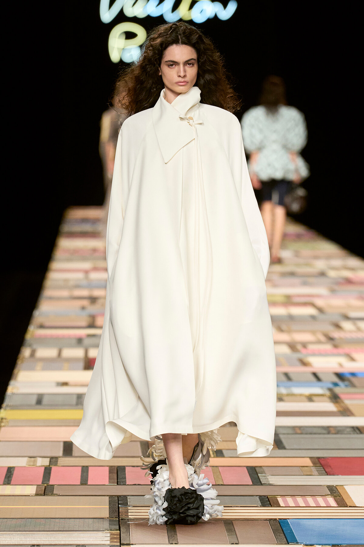 Model on a fashion runway in an off-white, cape-style coat with a high, asymmetrical collar and draped tie, walking on a colorful tiled catwalk.