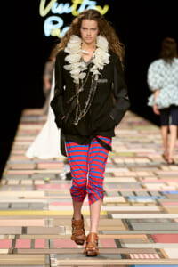 Model on a runway wearing a black blazer with a large white ruffled scarf, layered chains, red-and-blue striped cropped pants, and brown sandals.