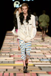 Model walks the runway in a cream blazer with a ruffled black scarf, layered necklaces, and blue-yellow striped pants with a front slit, black studded sandals.