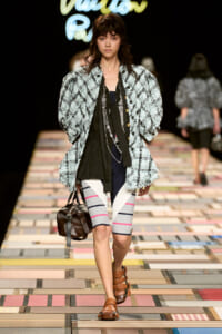 Model walks a fashion runway in a light-blue plaid textured coat over a black top, with gray striped leggings, brown sandals, and a brown handbag.